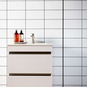 A wall with a white tile panel with black grout lines. A white vanity in front with soap and a wooden floor in the forefront.
