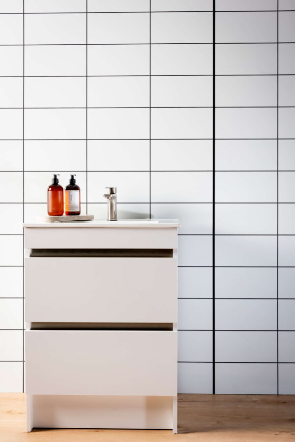 A wall with a white tile panel with black grout lines. A white vanity in front with soap and a wooden floor in the forefront.