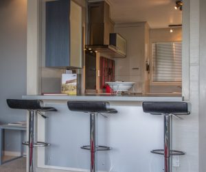 A kitchen breakfast bar with a silver wet wall kick plate underneath facing into a kitchen with three black bar stools, a bowl on the bar.
